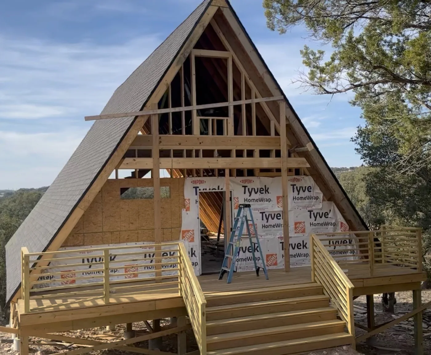 A-frame under construction, sheathed and wrapped in weather barrier, with a new deck across the front.